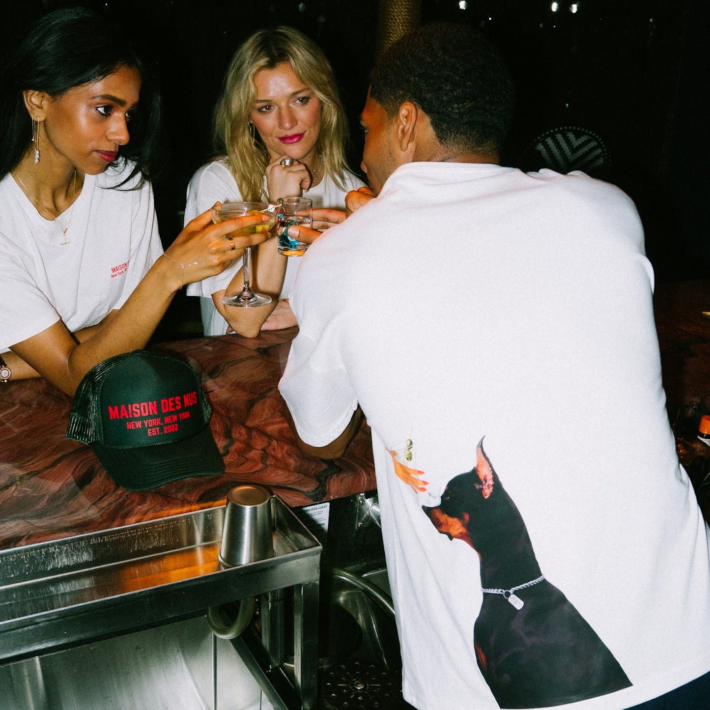 Three people at a bar counter with drinks, one wearing a white shirt with a dog graphic.