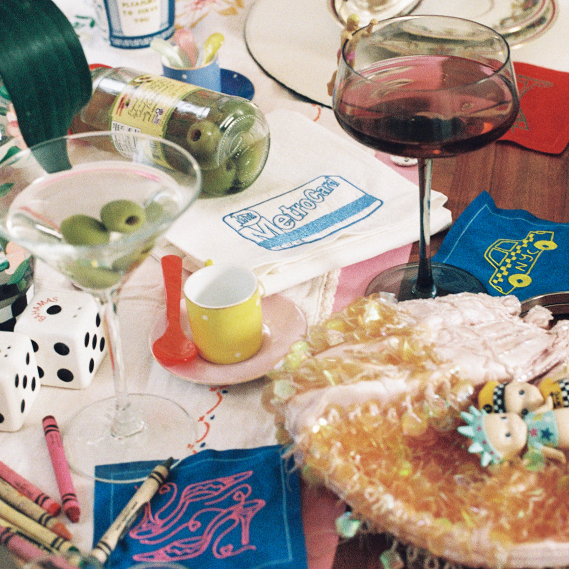 Messy table with party supplies, drinks, embroidered cocktail napkins and tea towels on a pink surface.